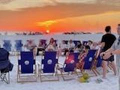 group of people sitting and standing around beach chairs on a sandy beach in a circle around an active bonfire with a sunsetting in the background