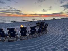 beach chairs on a sandy beach in a circle around an active bonfire with a sunset