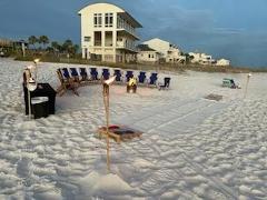 beach chairs on a sandy beach in a circle around a bonfire with tiki torches lit and a white house in the background