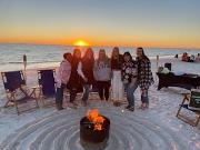 People standing on the beach in front of a bonfire wth the sun setting in the background.