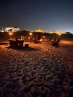 People sitting in beach chairs around a circle with a fire pit lit in the  center and tiki torches lit around the bonfire in the evening sandy beach.