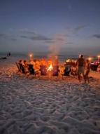 Photo of dark evening sky with  people sitting in beach chiars in a half circle on a sandy beach with a fire pit in the middle with a nice lit fire glowing in the night.