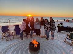 beach chairs on a sandy beach in a circle around a bonfire pit, group of people standing in the middle of the circle as the sun is setting over the water behind them